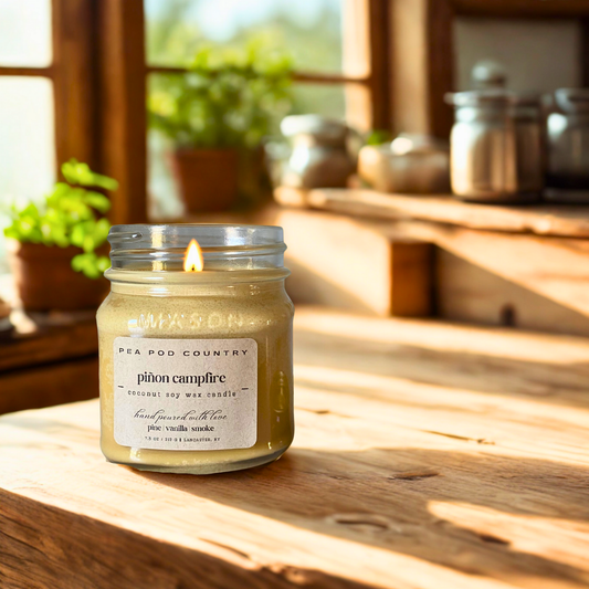 Candle in a jar labeled 'pinon campfire' on a wooden surface with a window and plants in the background